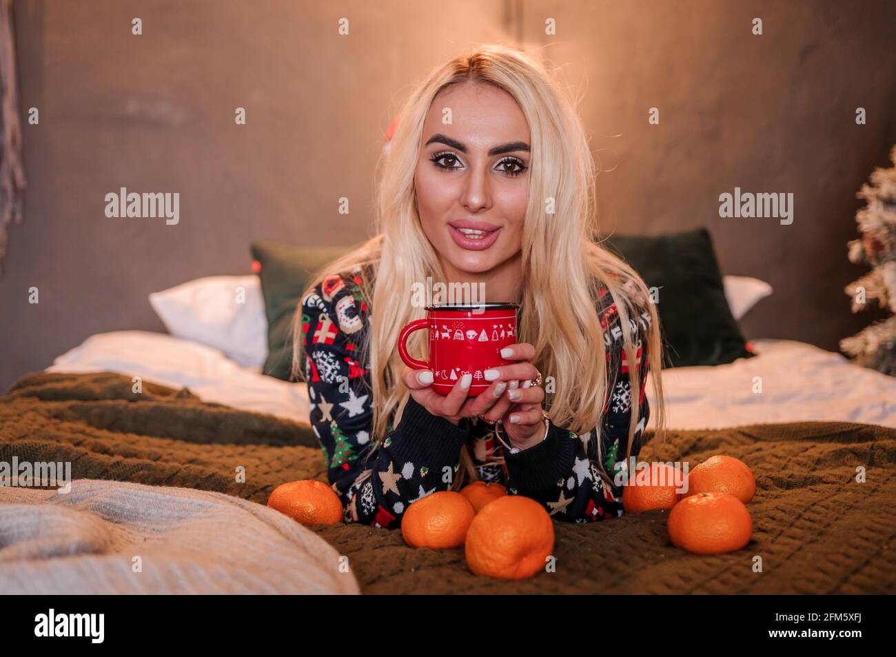 A girl in a sweater is lying on the bed. Tangerines on the bed and a red mug Stock Photo