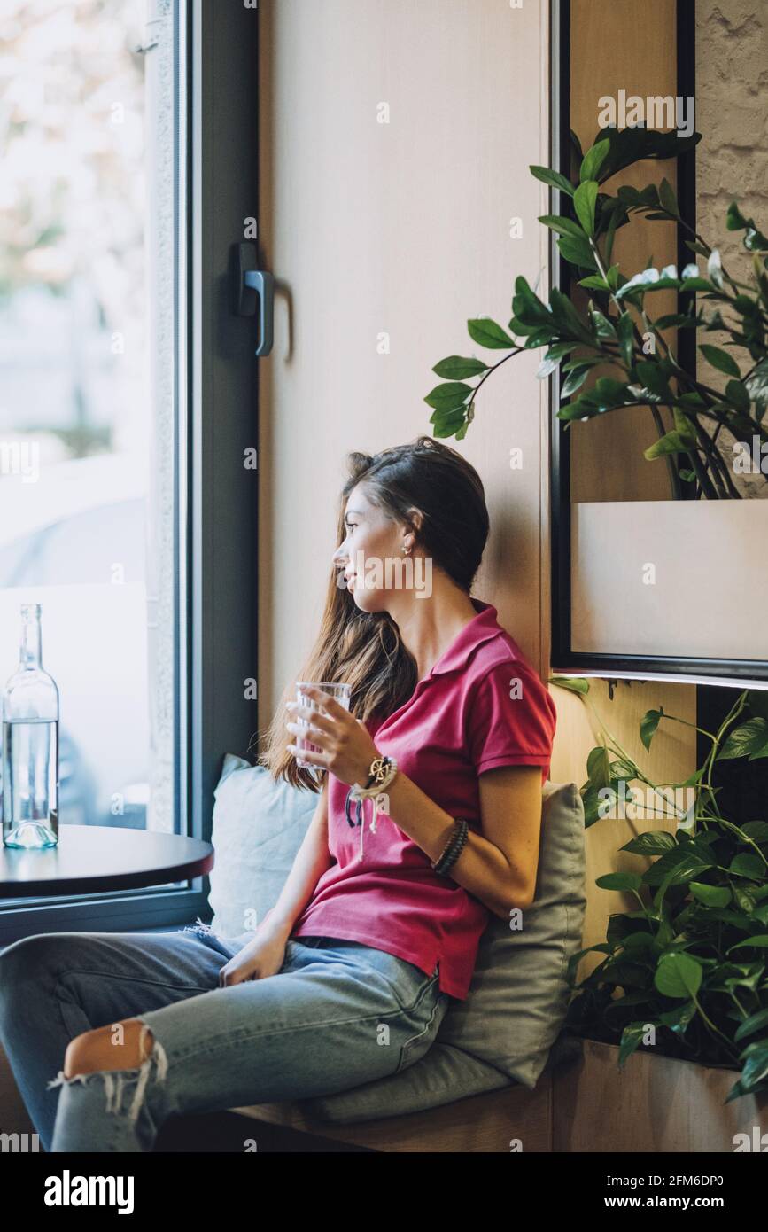Drinking more water, drink water reminder, keep water tracker. Brunette girl drinking water from a glass sitting in cafe by the window. Stock Photo