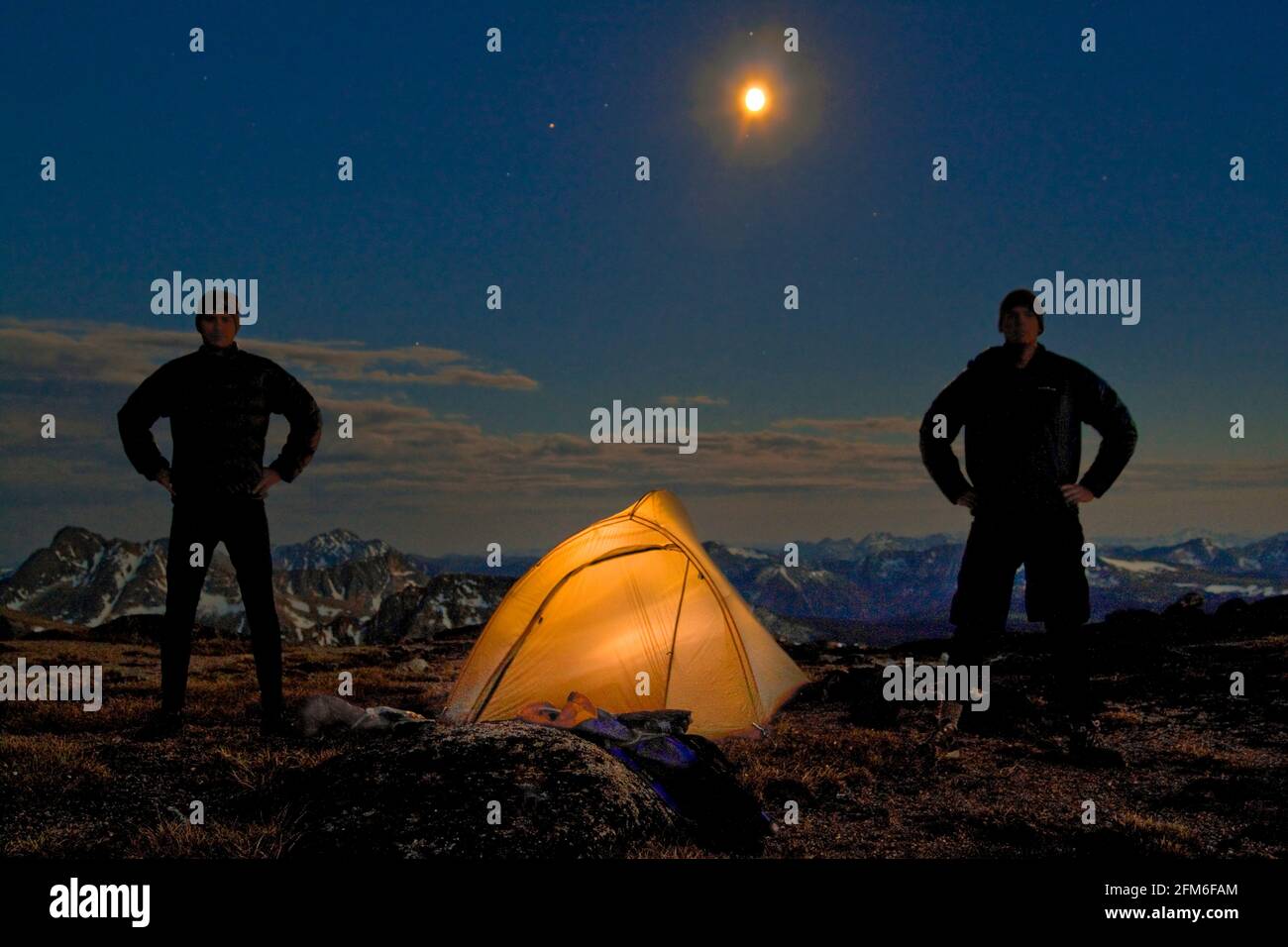 Two men stand next to their lit up tent on a mountain summit. Stock Photo