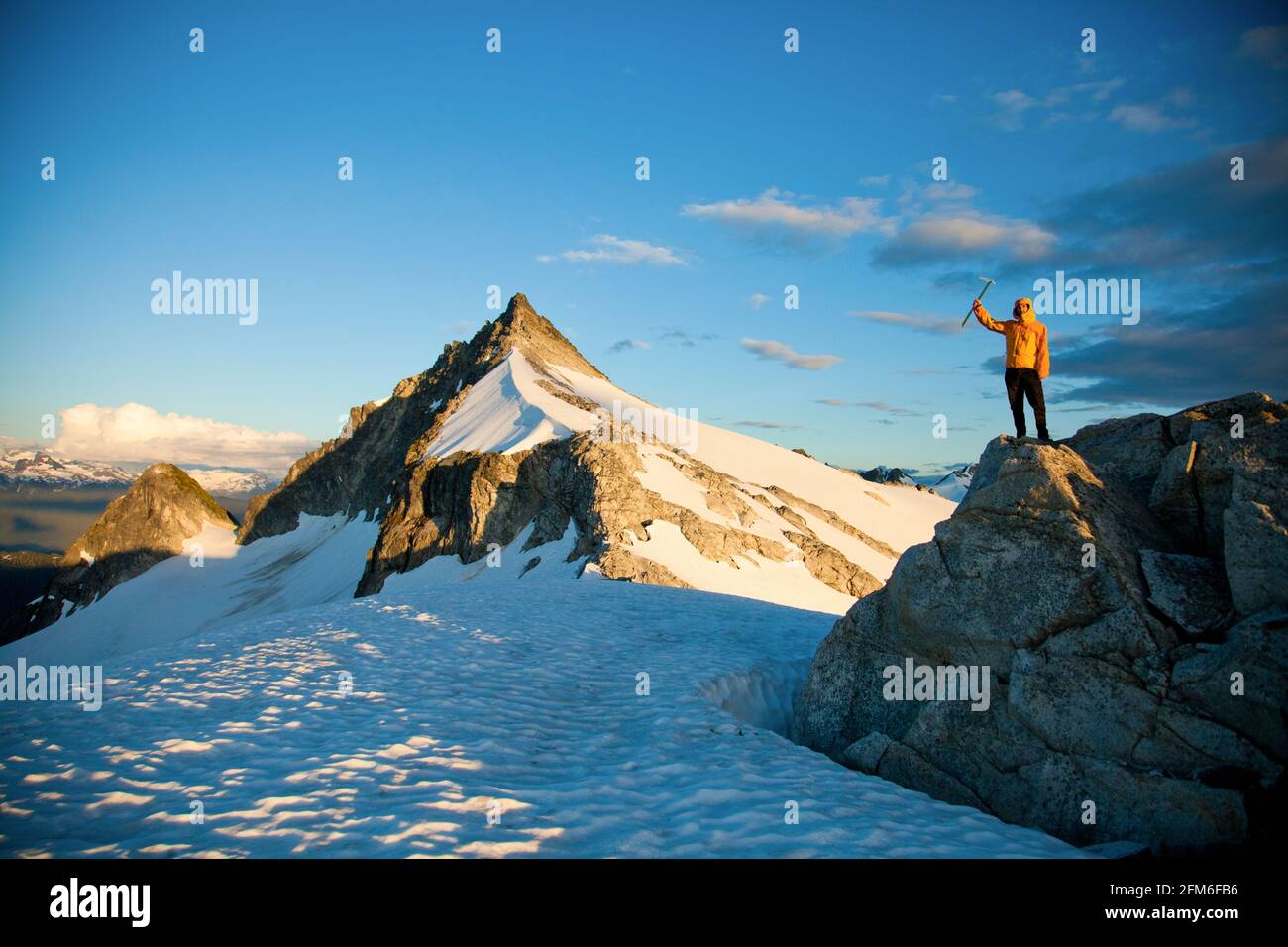 Successful climber wearing yellow jacket stands on rocky outcrop. Stock Photo