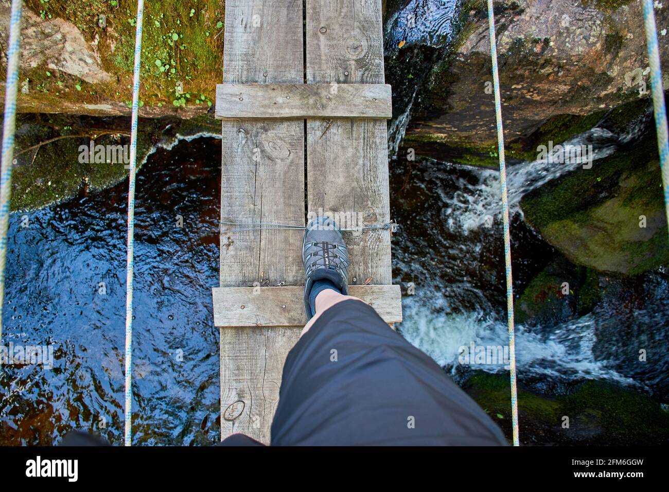 Leg of an athlete crossing a wooden bridge with ropes over a natural rock pool and waterfall. Stock Photo