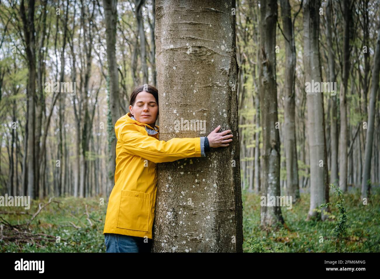 woman in yellow coat hugging a tree in the forest with eyes closed ...
