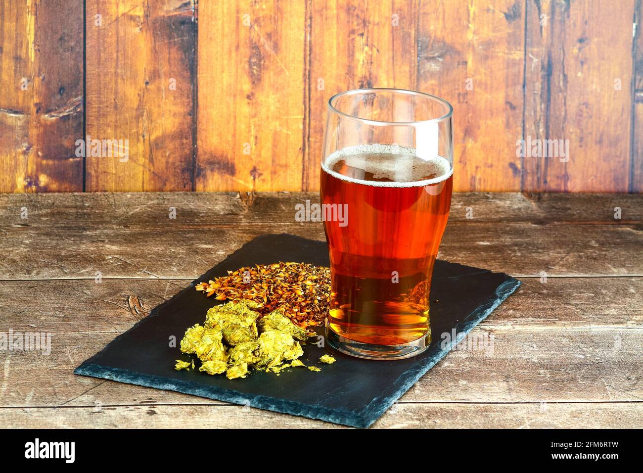 Pint glass filled with beer at the side of a slate filled with hops and brewers barley malt Stock Photo