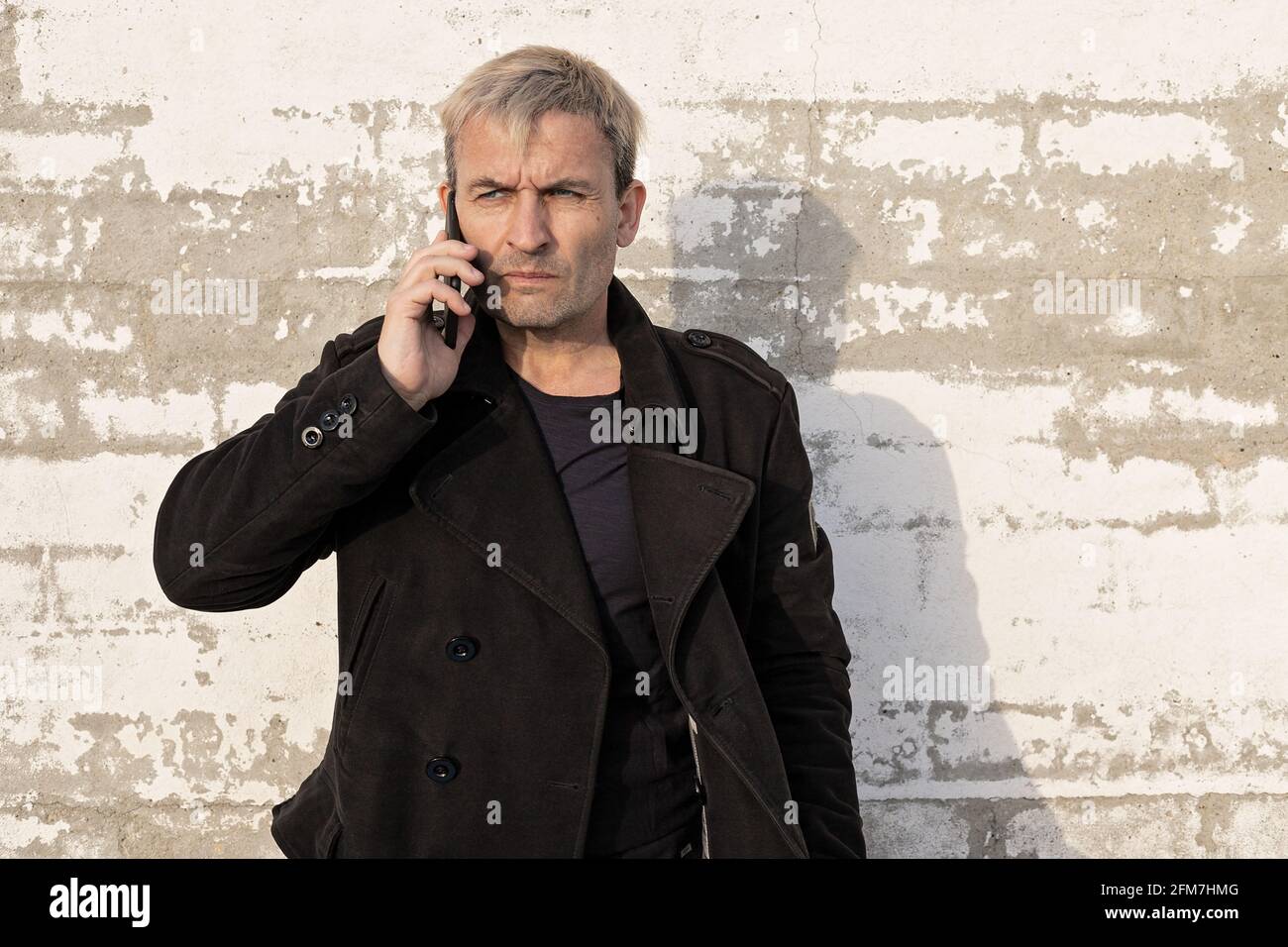 Attractive blonde middle aged man is posing with mobile phone against white brick wall. Male modelling, fashion photoshoot, professional. Front view. Stock Photo