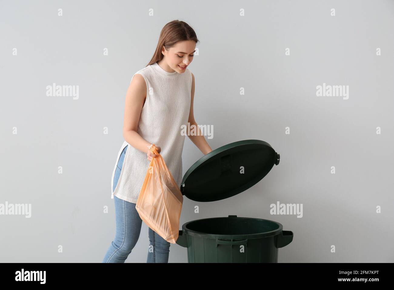 Young woman putting garbage in trash bin on light background Stock
