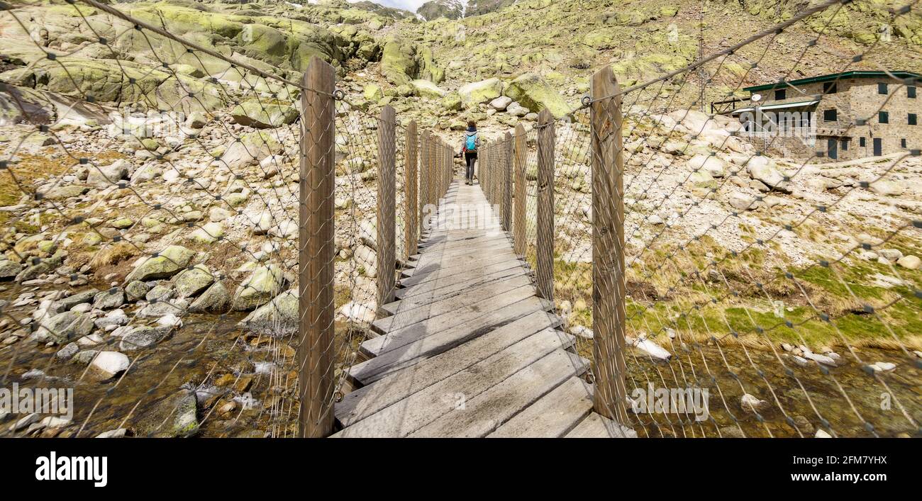 Young woman crossing a wooden bridge and hiking in the mountains. In the big lagoon of the Gredos circus, Avila in Castilla y Leon, Spain. Stock Photo