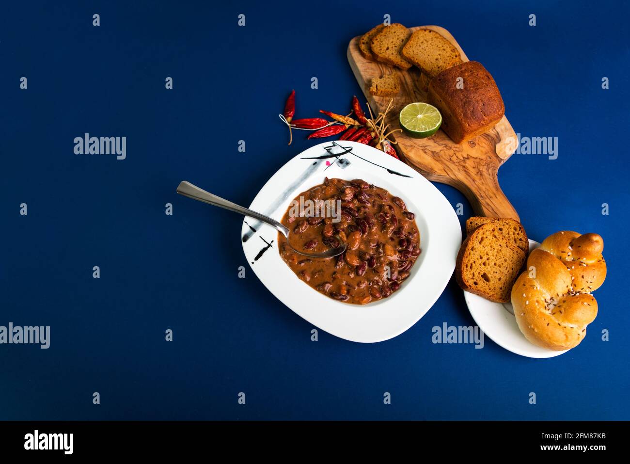 Bean soup in white plate, bread on kitchen board and plate, dried red peppers on blue background with big free space. Stock Photo
