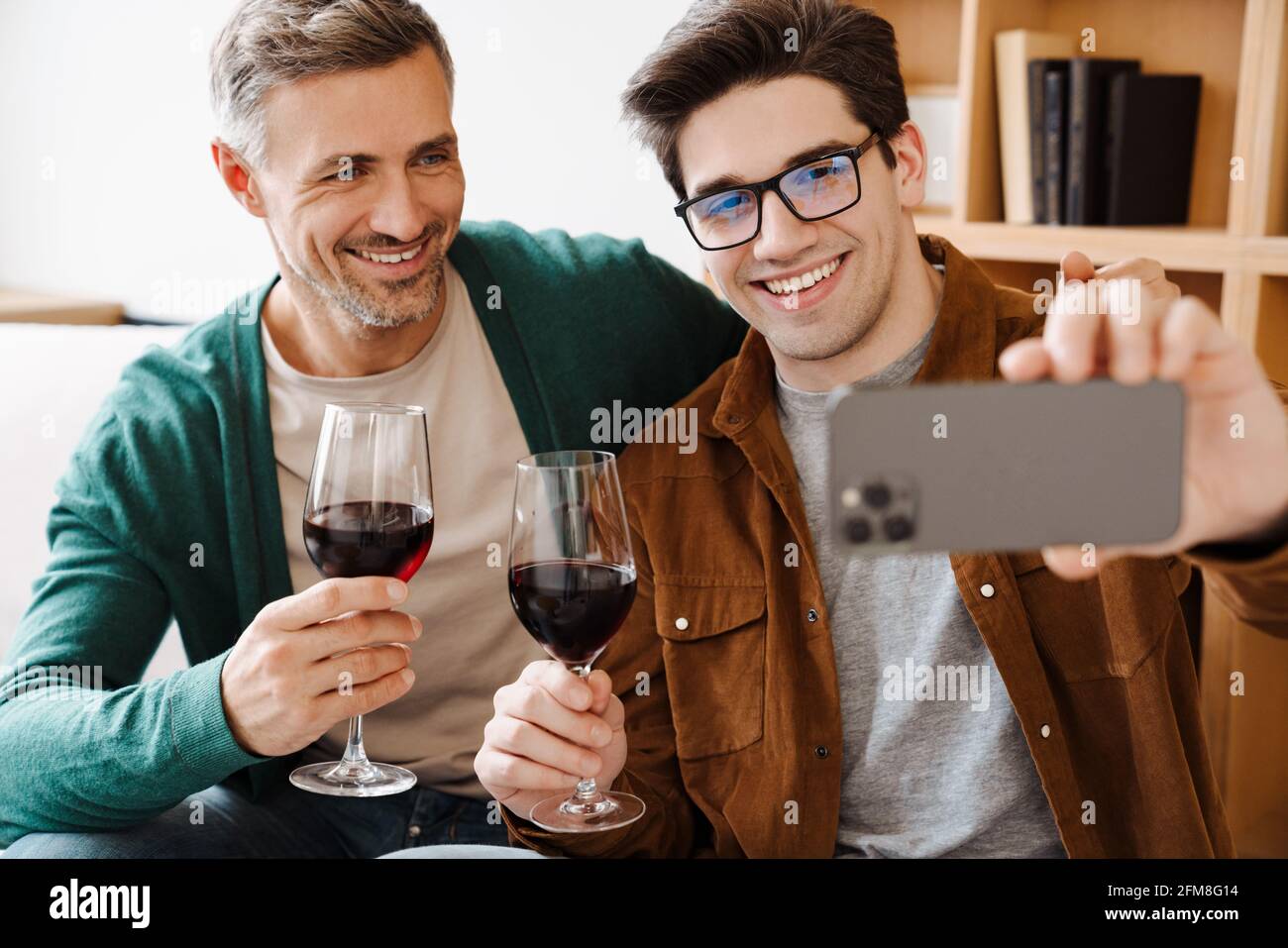 Happy gay couple hugging and drinking wine while taking selfie on cellphone at home Stock Photo