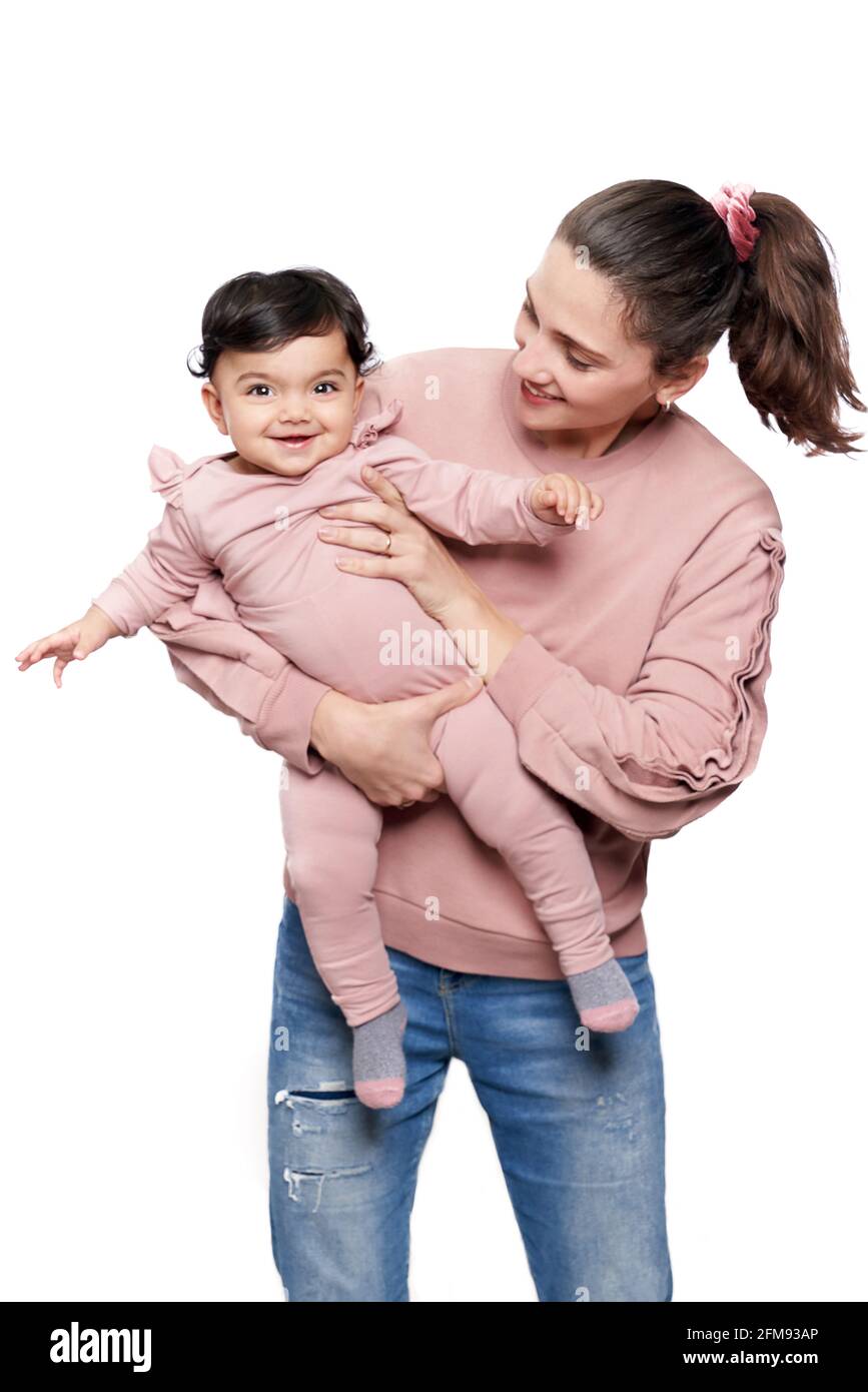Portrait of cute mother holding daughter isolated on white studio background. Front view of young attractive woman hugging sweet adorable smiling baby girl while posing and looking at camera. Stock Photo