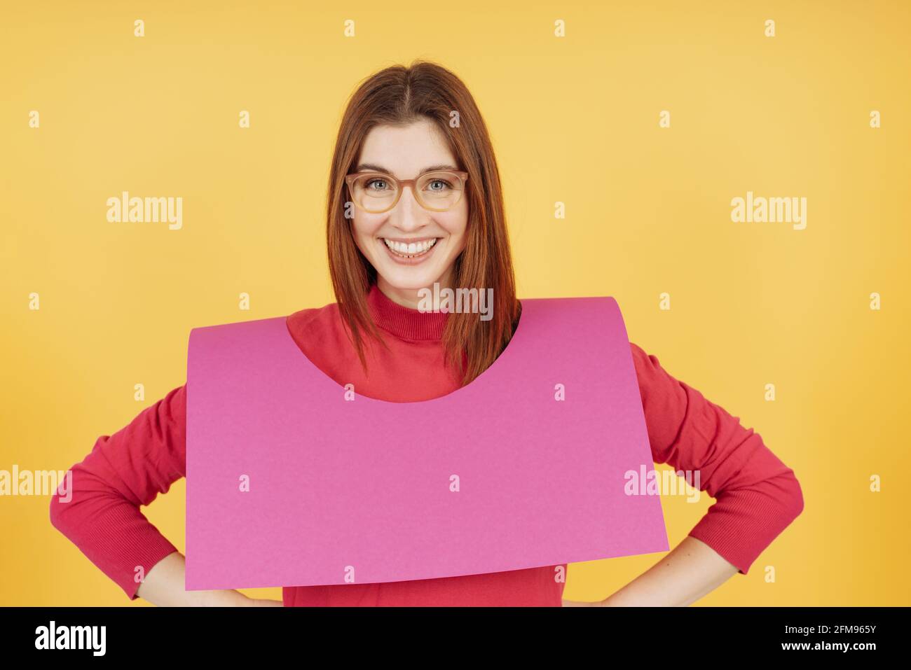 Vivacious laughing young woman wearing a pink card cutout draped over her shoulders as she grins at the camera with hands on hips over a yellow studio Stock Photo