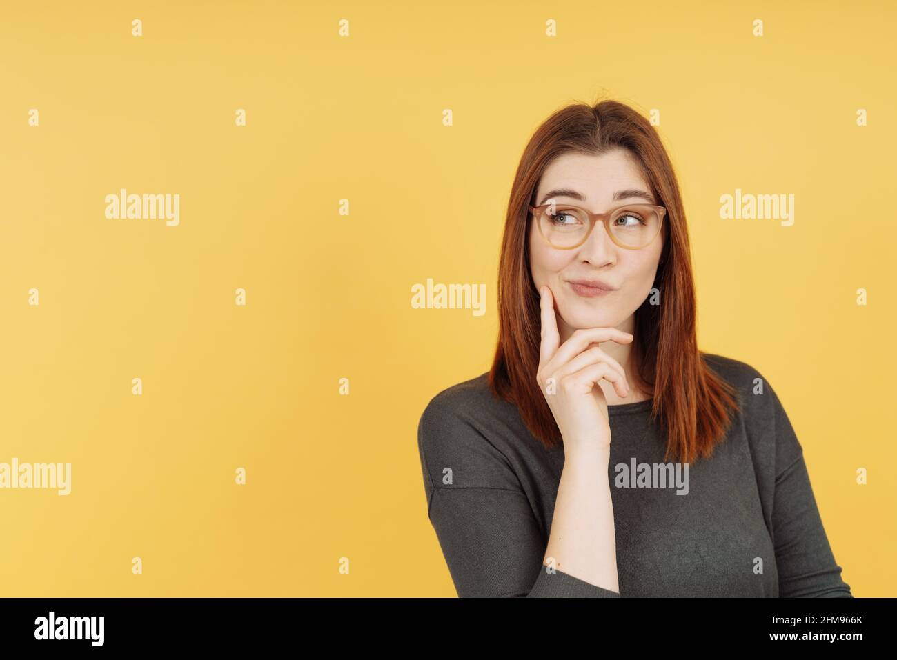 Pensive young woman mulling over a problem with a pensive smile and her hand to her chinas she looks aside with a wry expression over a yellow studio Stock Photo