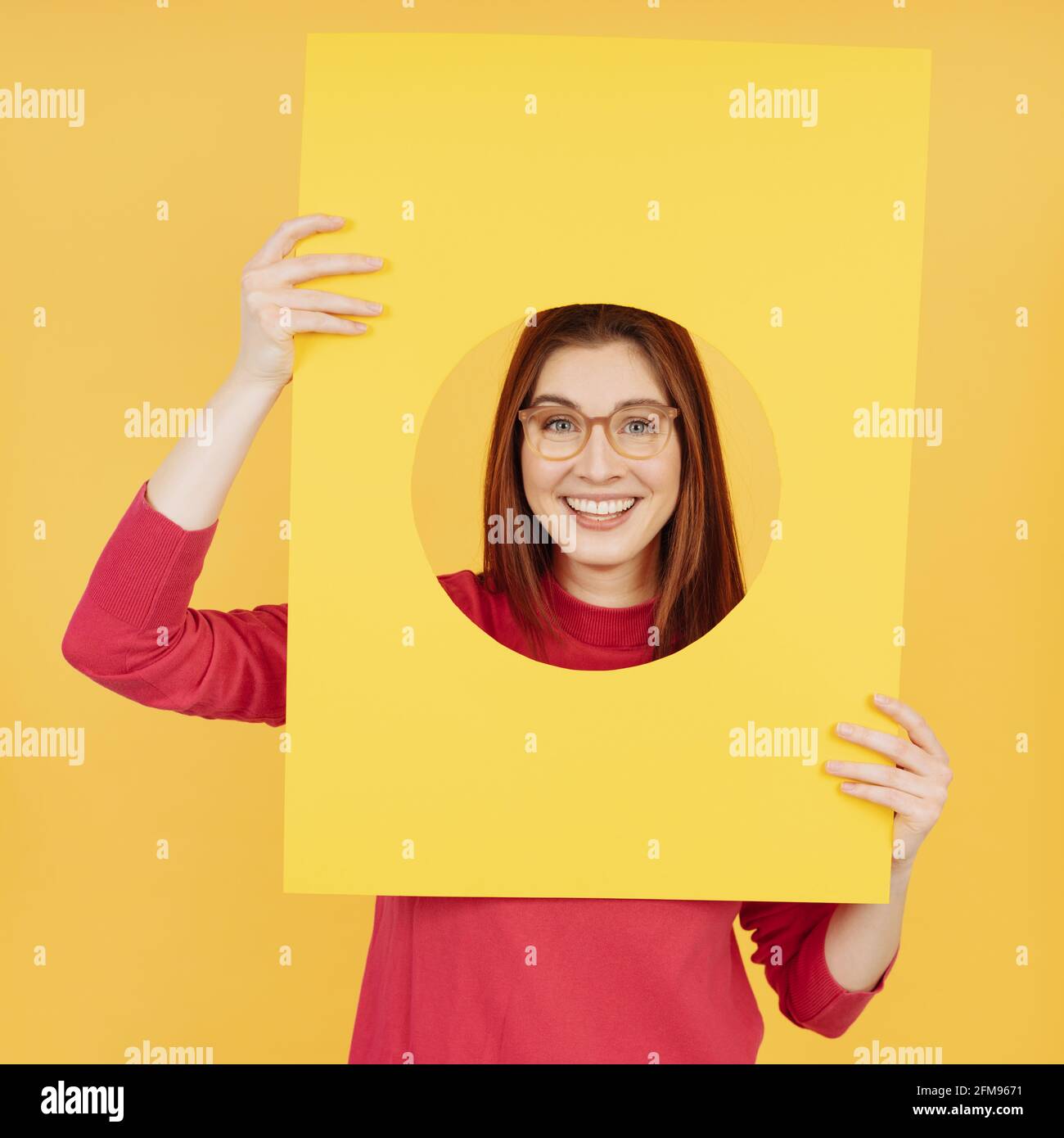 Fun playful young woman wearing glasses peering through a circular cutout in a yellow card at the camera with a mischievous smile over a matching yell Stock Photo