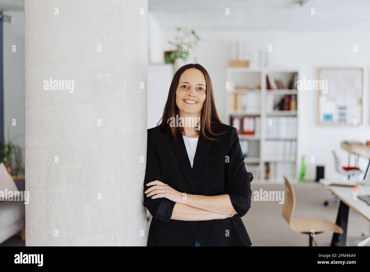 Relaxed businesswoman leaning on a pillar inside the office looking at the camera with a friendly smile and folded arms with copyspace on the side Stock Photo