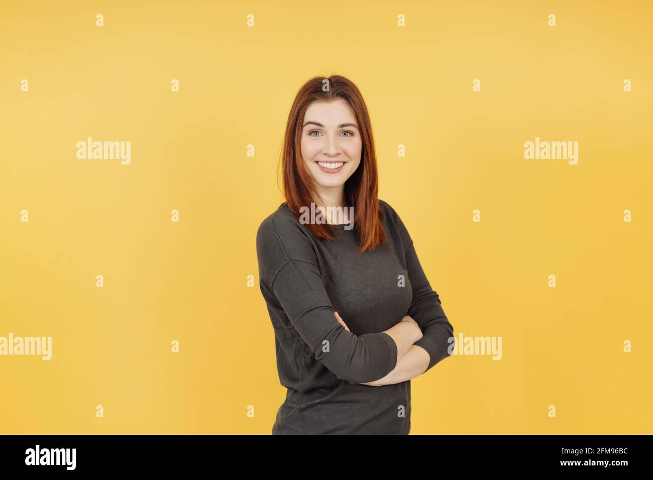 Attentive young woman looking at the camera with a warm friendly as she poses with folded arms over a yellow studio background with copyspace Stock Photo