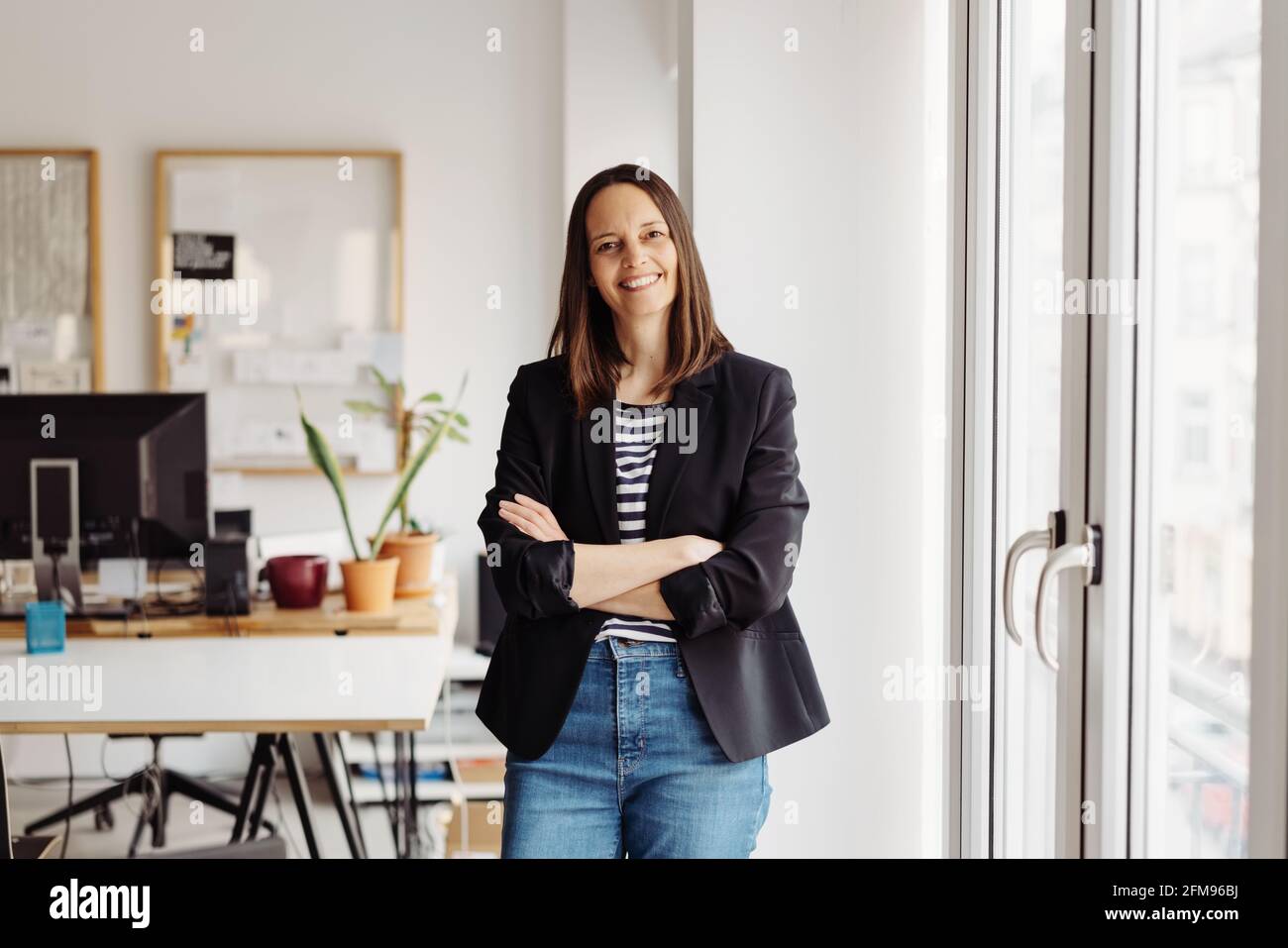 Smart modern businesswoman with a sincere kindly smile standing with folded arms near a high key glass door to the office with lateral copyspace Stock Photo