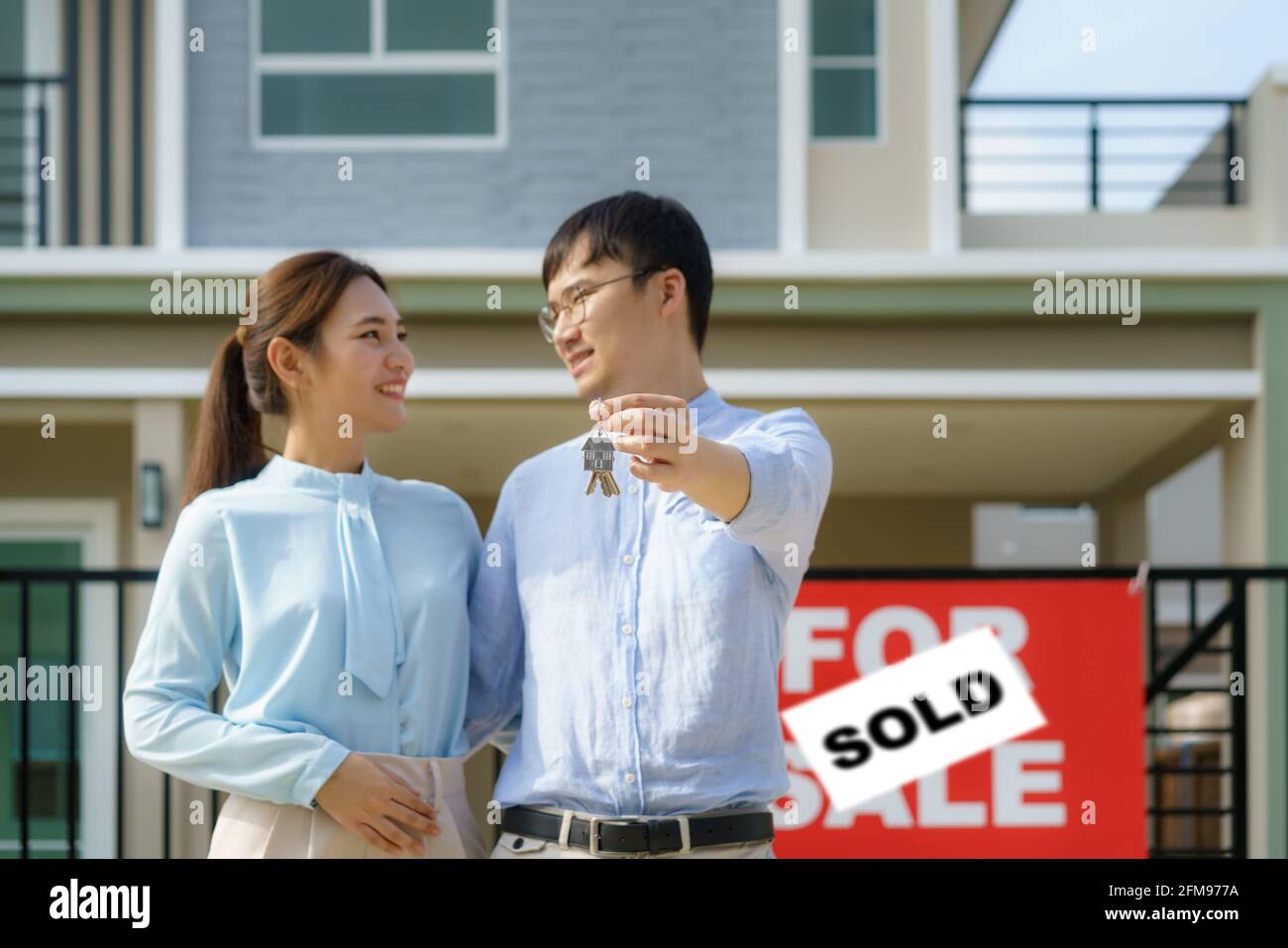 Portrait of Asian young couple standing and hugging together and holding house key looking happy in front of their new house to start new life. Family Stock Photo