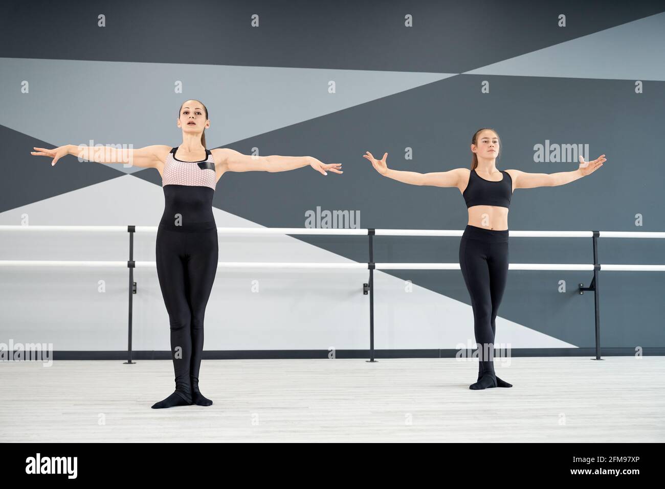 Adult female instructor helping young girl in black sportswear learn choreographic move. Two synchronized women practicing in hall, hi tech interior. Choreography, gymnastics concept. Stock Photo
