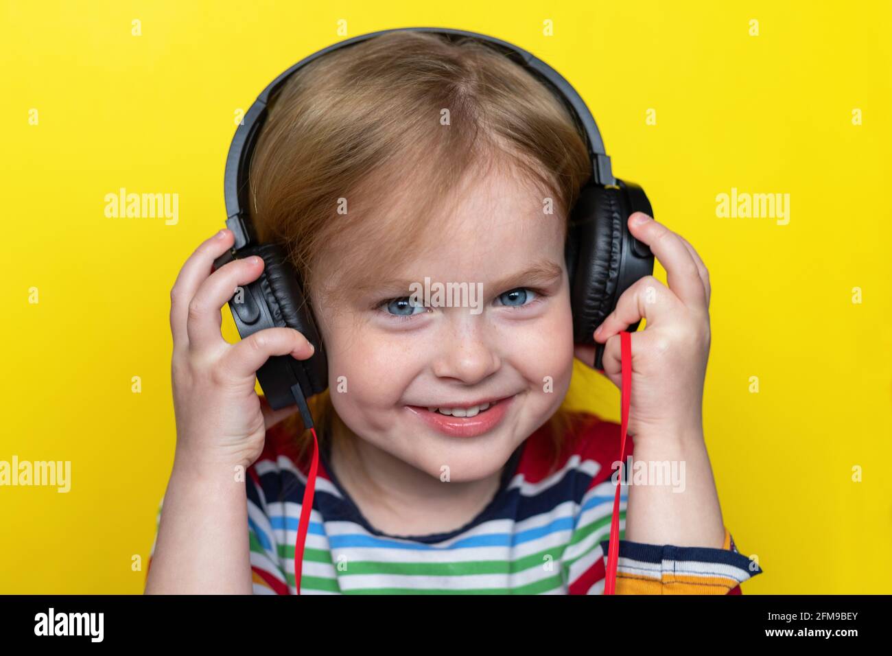 Happy smiling little child listening to music with large headphones, isolated on yellow background. Stock Photo