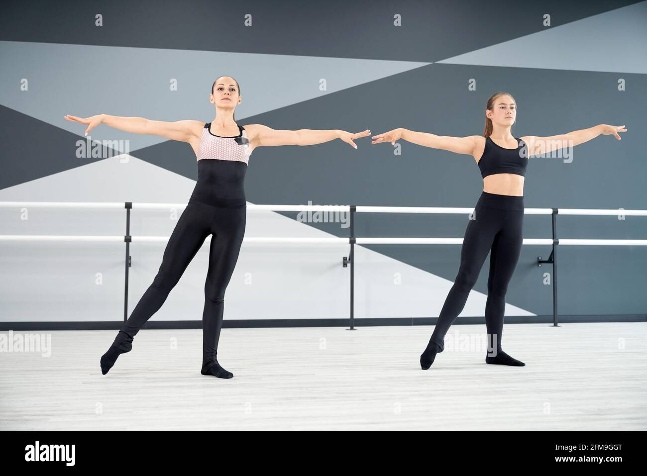 Adult female instructor helping young girl in black sportswear learn choreographic move. Two synchronized women practicing in hall, hi tech interior. Choreography, gymnastics concept. Stock Photo