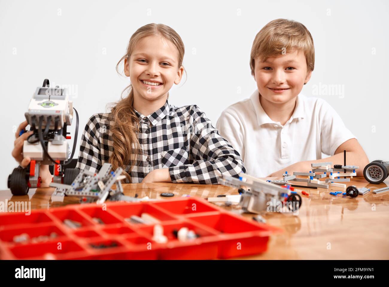 Front view of boy and girl creating robot, red box with building kit on table. Nice interested friends smiling, lookig at camera and working on project together. Concept of science engineering. Stock Photo