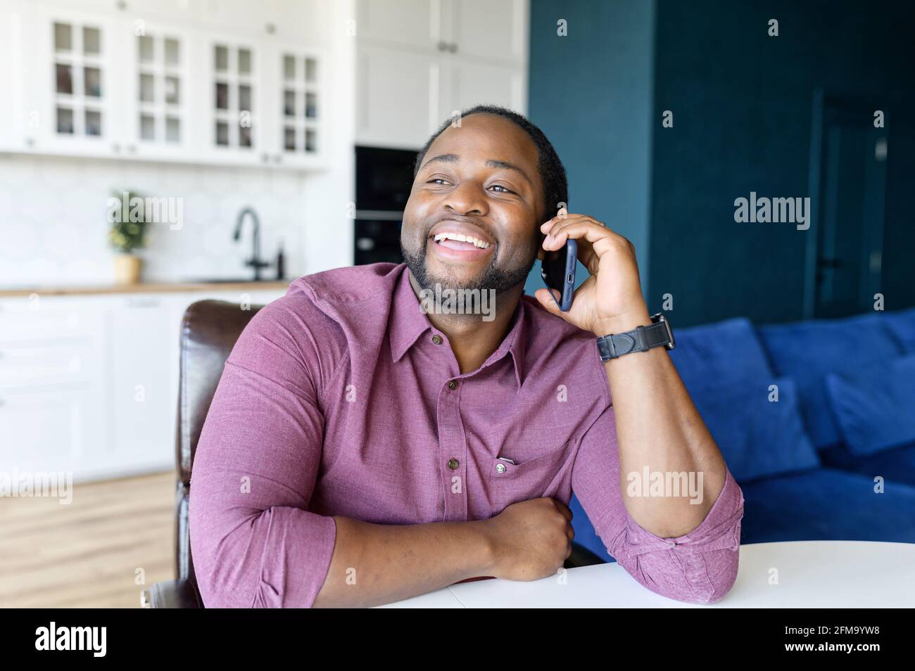 Glad to hear you. Happy African-American guy enjoy a phone conversation with friend or family, talking on the mobile phone sitting at the desk in cozy living room, black guy speaking on the cellphone Stock Photo