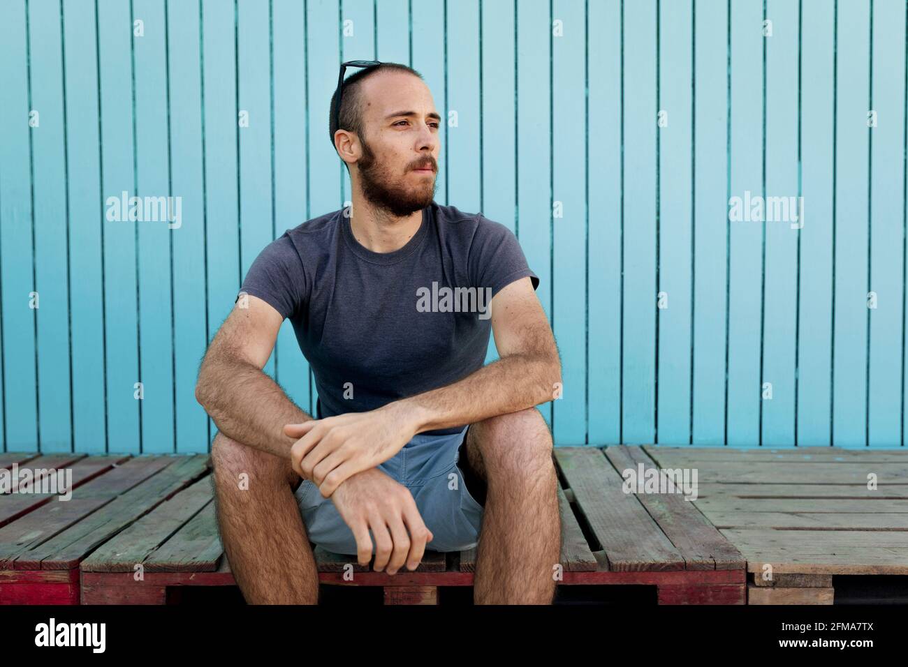 Bearded young man wearing sunglasses over the head is confidently looking away at the Isla Canela beach with a blue wooden wall background Stock Photo