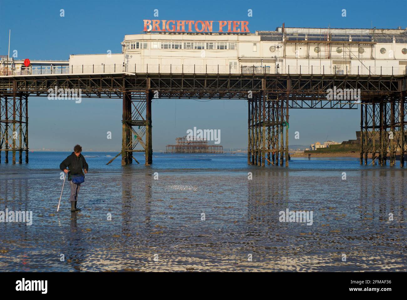 Brighton's dilapidated West Pier framed in the structure of 'Brighton Pier', the Palace Pier. Fisherman collecting ragworm for bait in the foreground. Stock Photo
