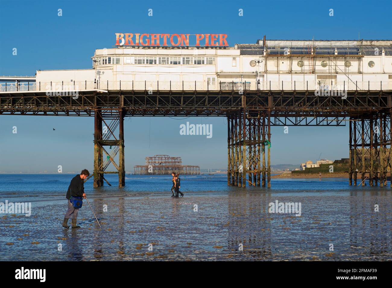 Brighton's dilapidated West Pier framed in the structure of 'Brighton Pier', the Palace Pier. Fisherman collecting ragworm for bait in the foreground, women going for a swim behind. Stock Photo
