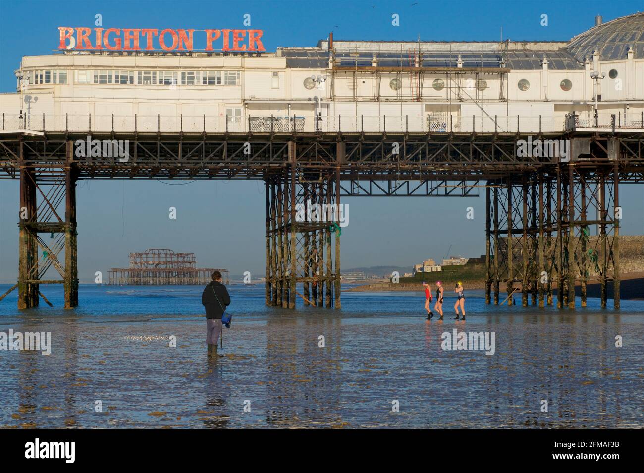 Brighton's dilapidated West Pier framed in the structure of 'Brighton Pier', the Palace Pier. Fisherman collecting ragworm for bait in the foreground, women going for a swim behind. Stock Photo