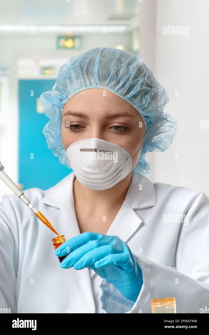 Pharma female tech works in laboratory. Caucasian young woman in protective gloves, face mask, hat and white gown pipettes sample in tube. Lab Stock Photo