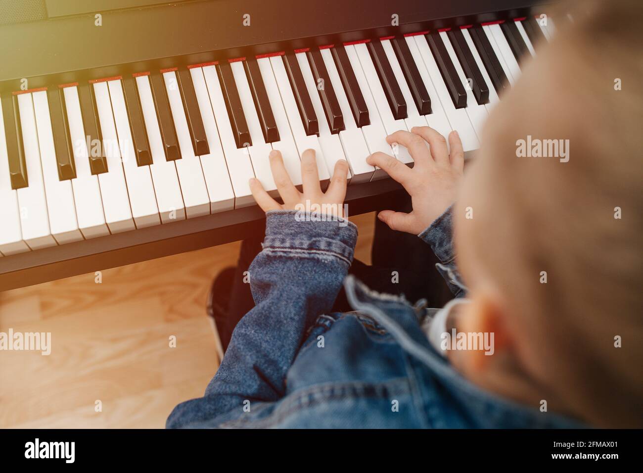 Little boy playing electric piano at home, sun is hitting keyboard. He is wearing jeans jacket, his blond hair in a bun. Blurred, high angle, close up Stock Photo