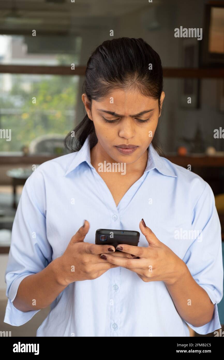 Vertical shot of a worried young Indian female holding her phone with two hands and looking at it Stock Photo