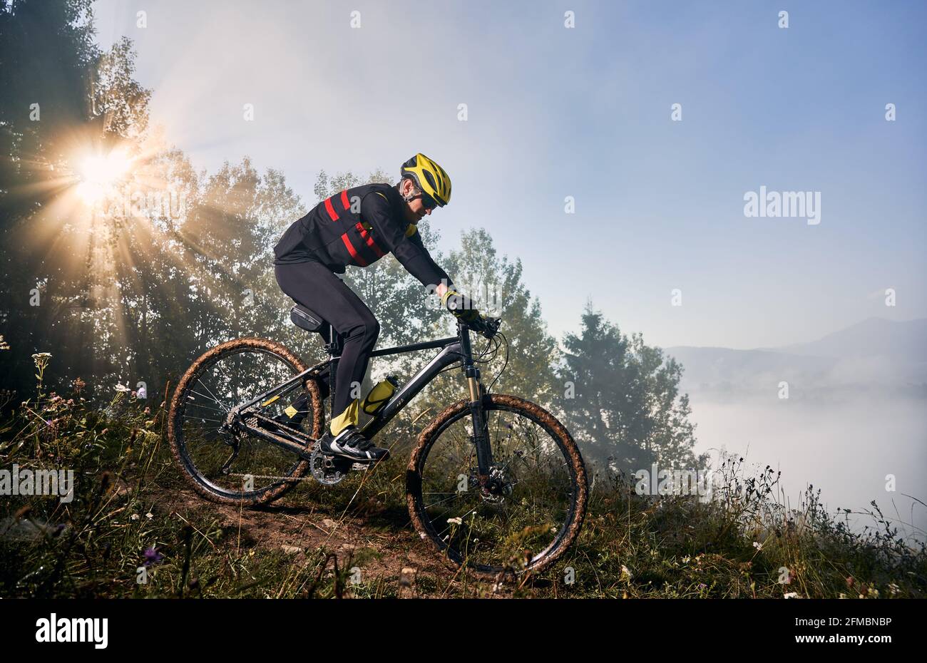 Horizontal snapshot of man riding his bike in the mountains in early foggy morning going downhill. Sun is shining through tree crown, peaks in clouds on background. Concept of active lifestyle Stock Photo