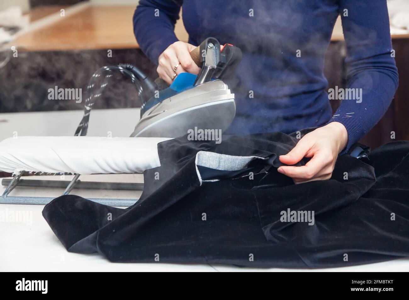 Seamstress steams a velvet jacket with an iron in her atelier. Authentic lifestyle. Stock Photo