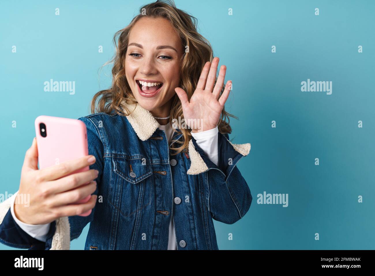 Blonde smiling woman gesturing while using mobile phone isolated over blue wall Stock Photo