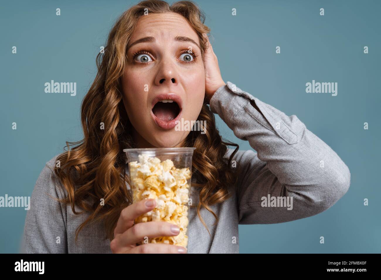 Shocked young woman watching TV and eating popcorn isolated over blue background, scary movie