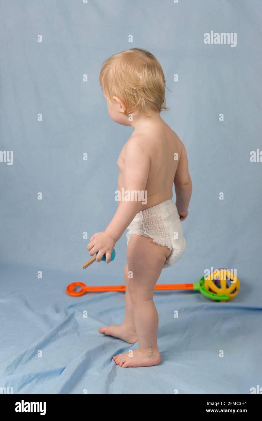 A small blond boy of 1 year in a white diaper stands on a blue background. View from the back Stock Photo