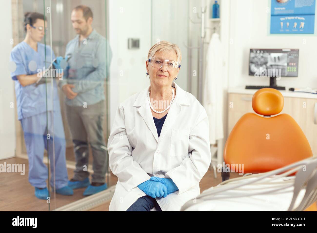 Senior stomatological woman looking into camera while sitting on dental chair in hospital stomatology room. Medical nurse discussing with patient during writing healdcare treatment Stock Photo