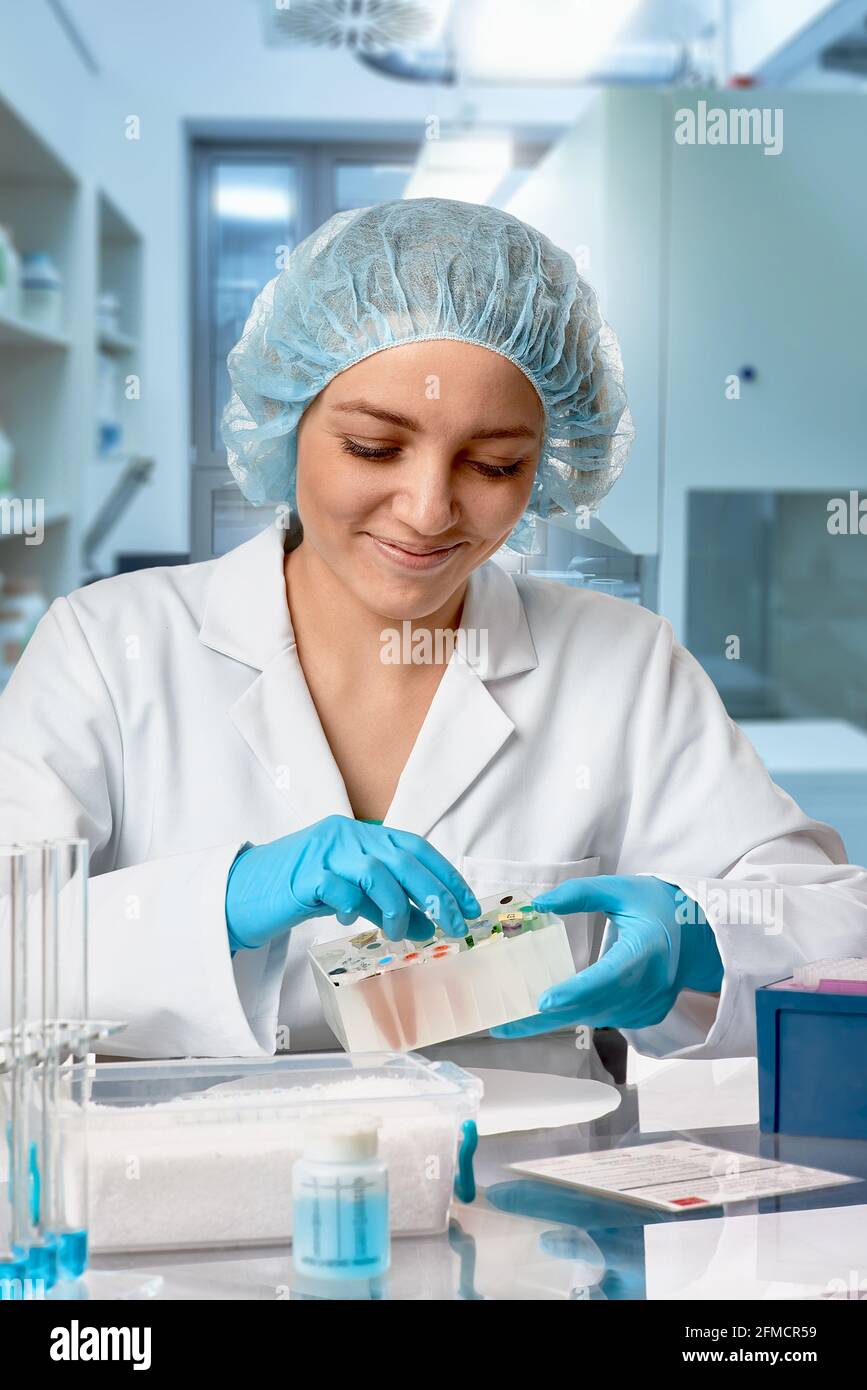 Pharma female tech works in laboratory. Caucasian young woman in protective gloves, hat and white gown handles sample tubes. Lab interior, selective Stock Photo