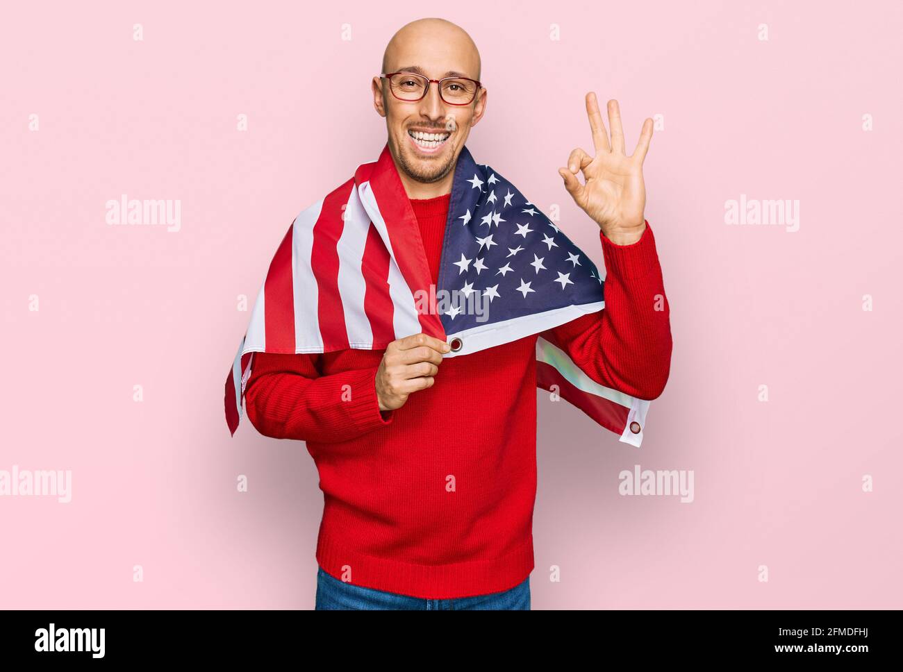 Bald man with beard wrapped around united states flag doing ok sign with fingers, smiling friendly gesturing excellent symbol Stock Photo