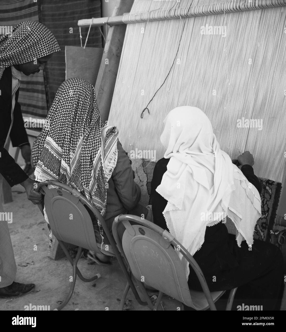 1960s, historical, two male Bedouin arabs in traditional dress and headwear, one wearing a tablecloth-like pattern, the other a plain white cloth, sitting outside doing traditional weaving, making a rug on a hand-loom, Jeddah, Saudi Arabia. The discovery of oil and the industrialisation of the country saw traditional handcrafts such as weaving go to decline. Stock Photo