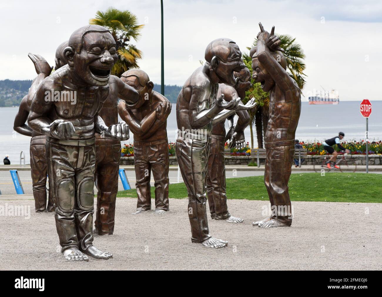 A cyclist rides past the statues from the sculpture A-Maze-ing Laughter by artist Yue Minjun stand along English Bay, where a freighter is anchored, Stock Photo