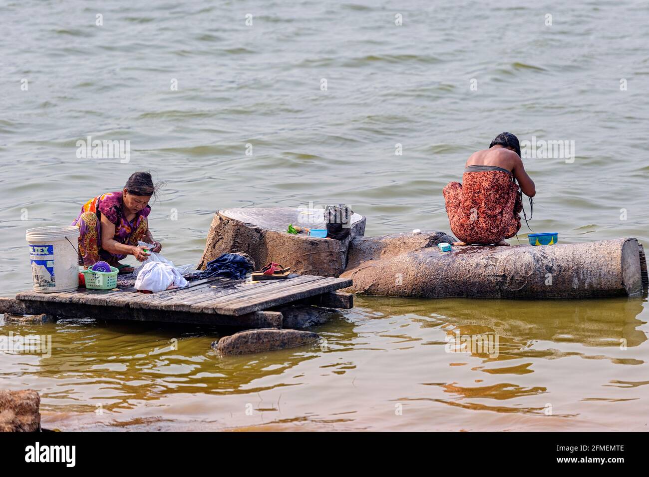 Two women wash clothes by hand on rocks and wooden planks along the edge of a calm  on Pindaya Lake. Pindalay Myanmar Burma Stock Photo