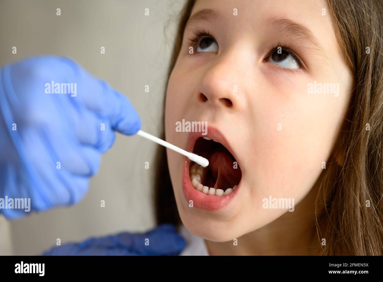COVID-19 PCR test and kid face close-up, little girl opens mouth for coronavirus testing. Doctor or nurse holds swab for saliva sample from cute child Stock Photo