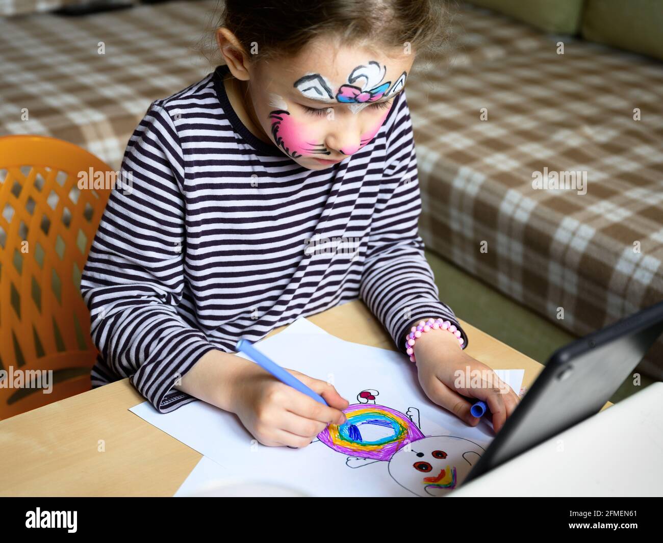 Kid drawing indoor, little girl with painted mask on face studying at home. Cute child learns to draw at table in room. Preschooler and color picture Stock Photo