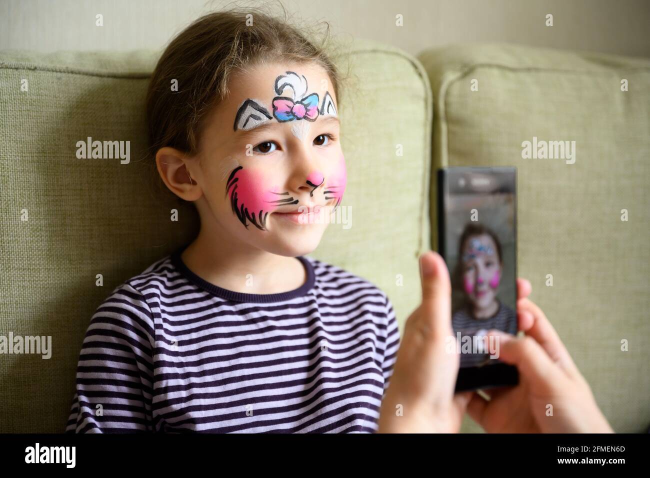 Kid with face painting of kitty, cute little girl with painted mask on face poses for photo. Happy pretty child with beautiful makeup as drawing. Ador Stock Photo