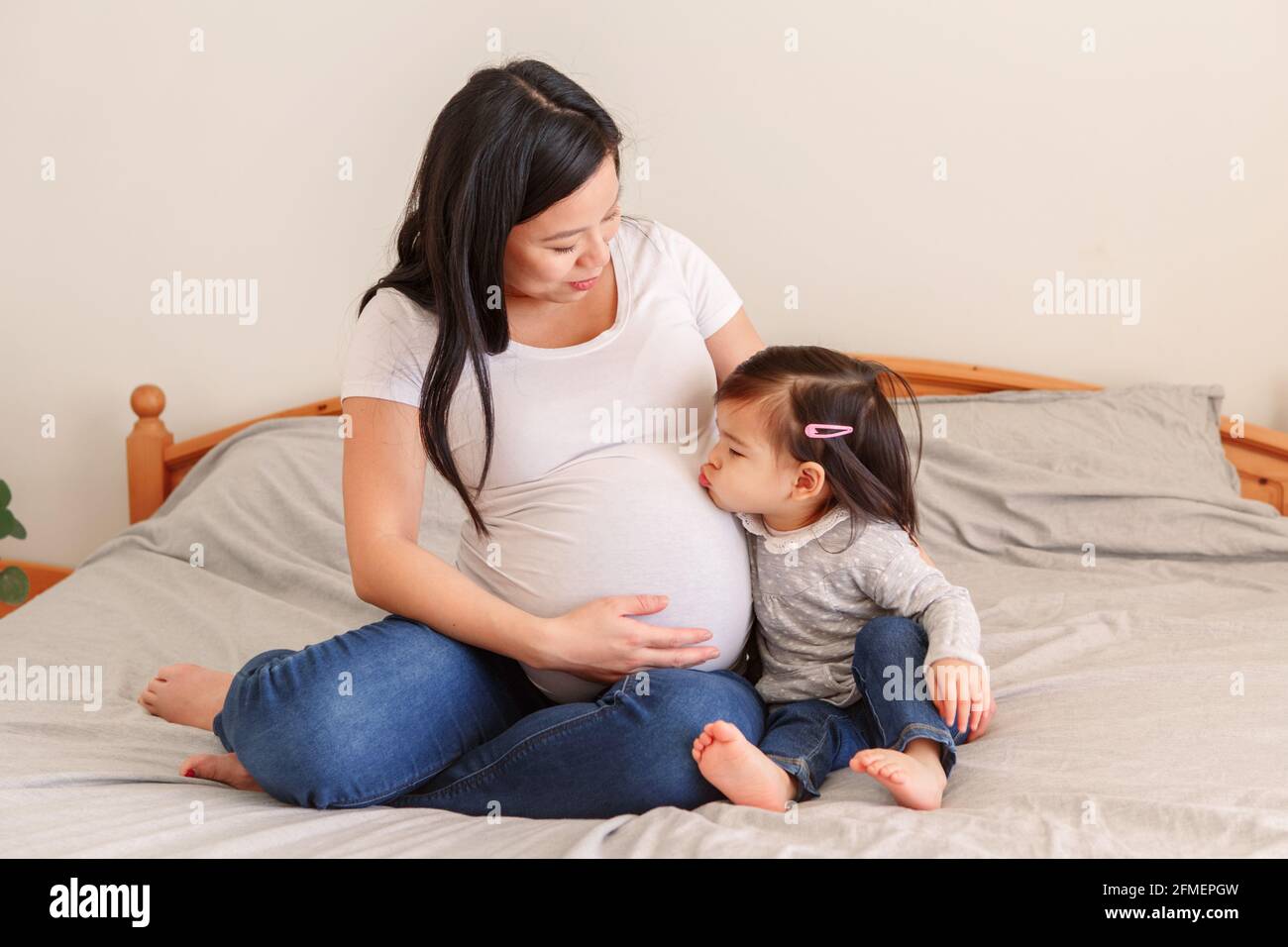 Asian Chinese Pregnant Woman With Toddler Girl Sitting On Bed At Home asian-chinese-pregnant-woman-with-toddler-girl-sitting-on-bed-at-home