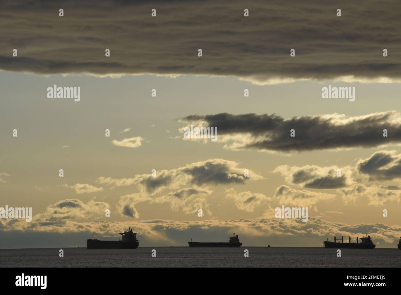 Silhouetted freighters ships are anchored in English Bay offshore under clouds lit by the setting sun in Vancouver, British Columbia, Canada Stock Photo