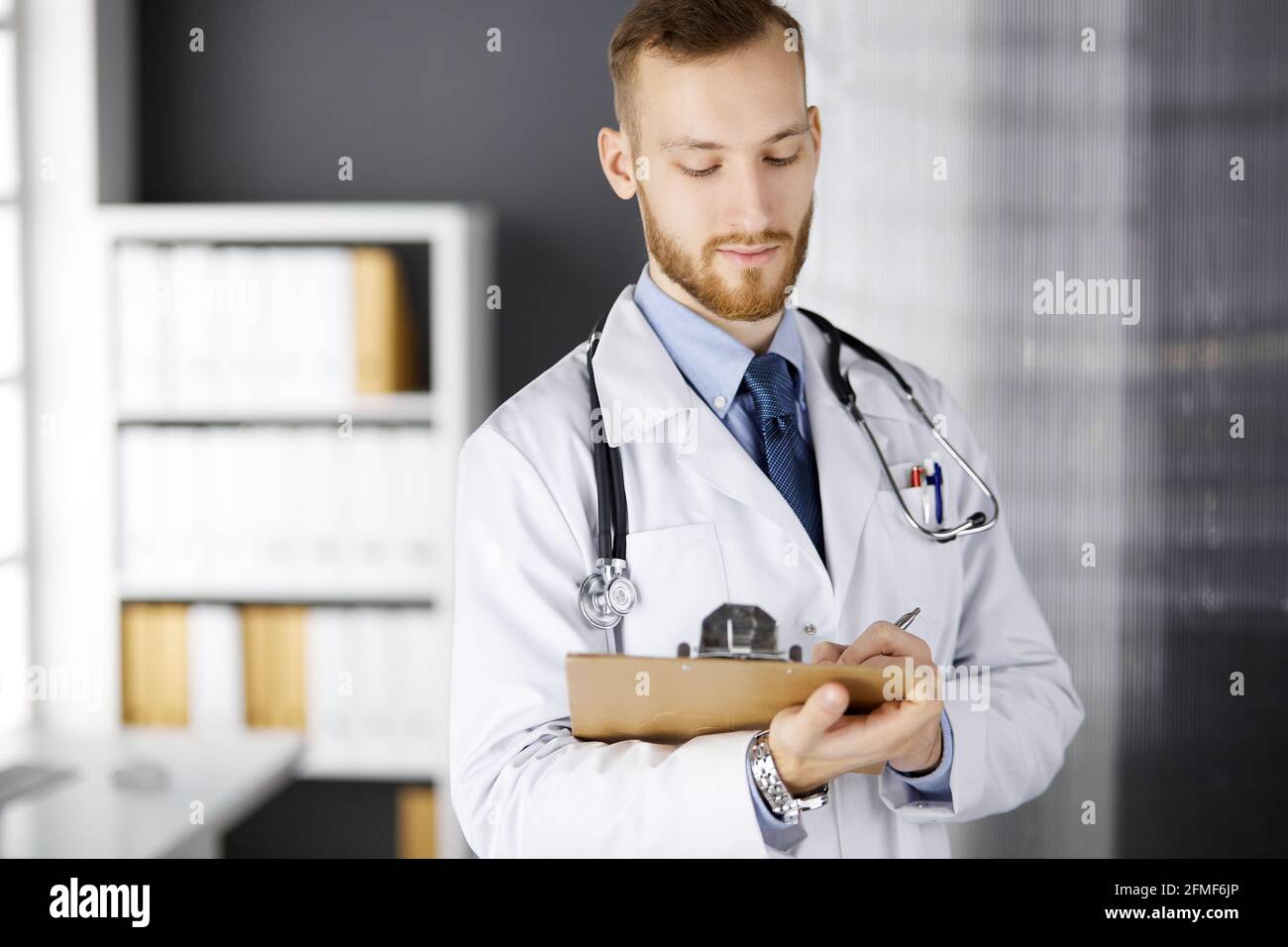 Friendly red-bearded doctor standing and writing with clipboard in ...