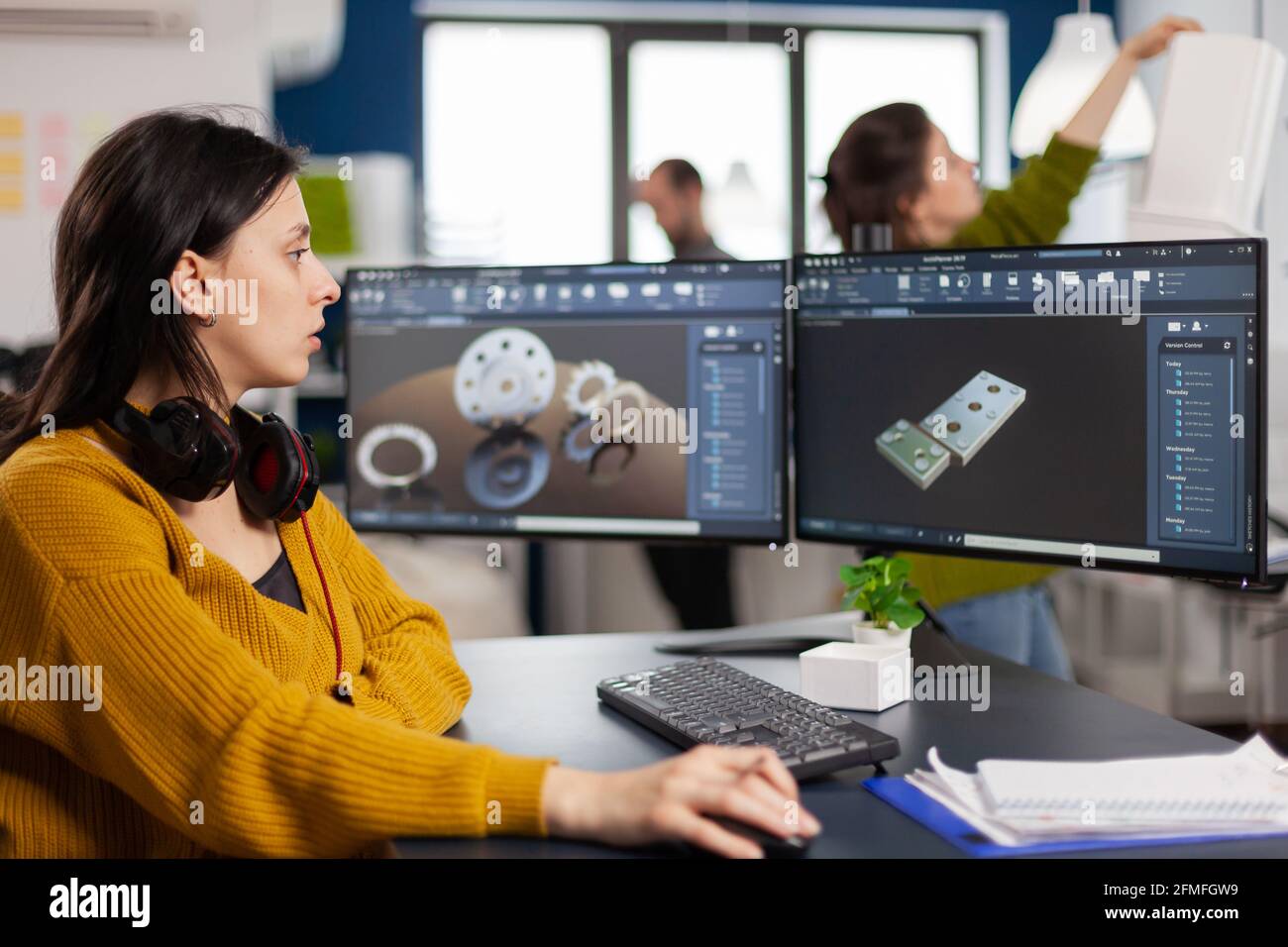 Industrial female engineer looking at personal computer with dual monitors setup, screens showing CAD software with 3D prototype of gears metalic mechanical piece. Employee working in creative office Stock Photo